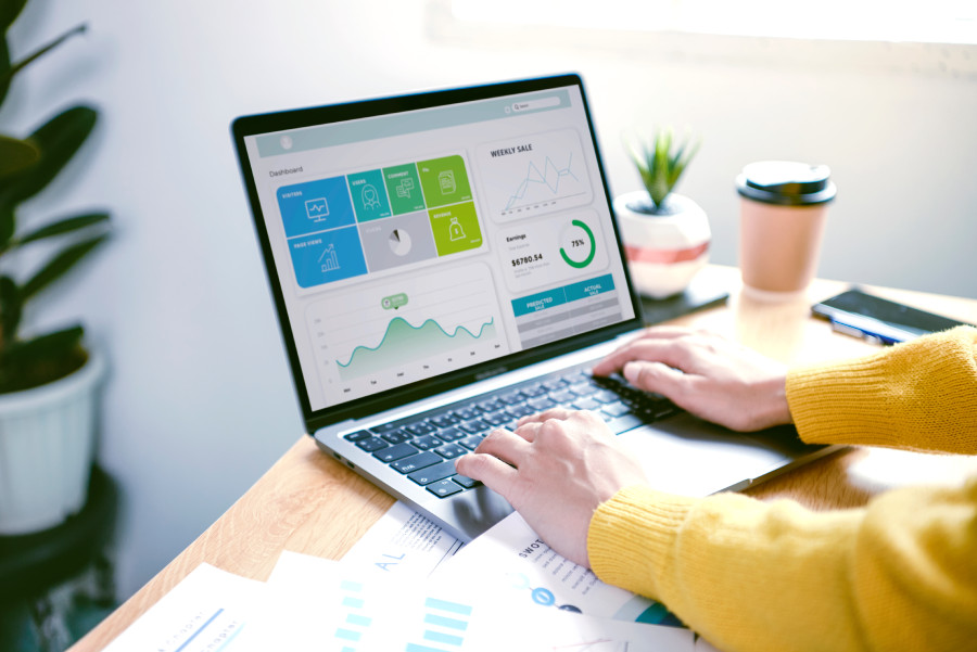 A person wearing a yellow sweater is working on a laptop displaying a dashboard for a B2B SaaS application. The screen shows various metrics, including weekly sales, a line graph, and numerical data. The workspace includes a small potted plant, a takeaway cup, and scattered papers.